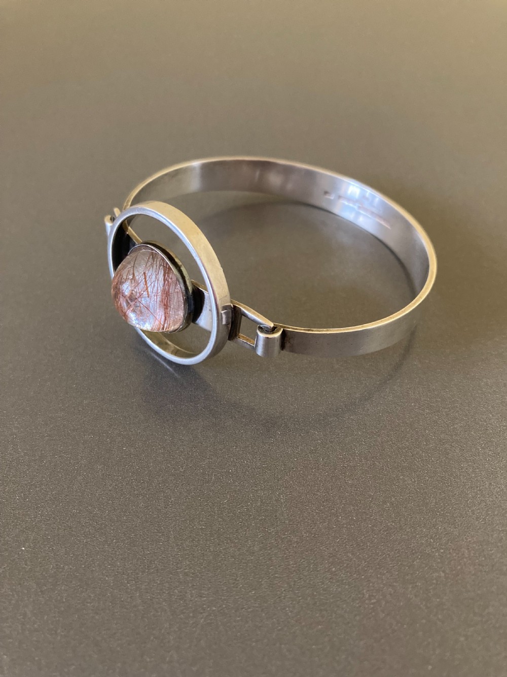 A hinged silver bangle features a central circular frame holding a cabochon rutilated quartz gemstone with needle-like inclusions. View II
