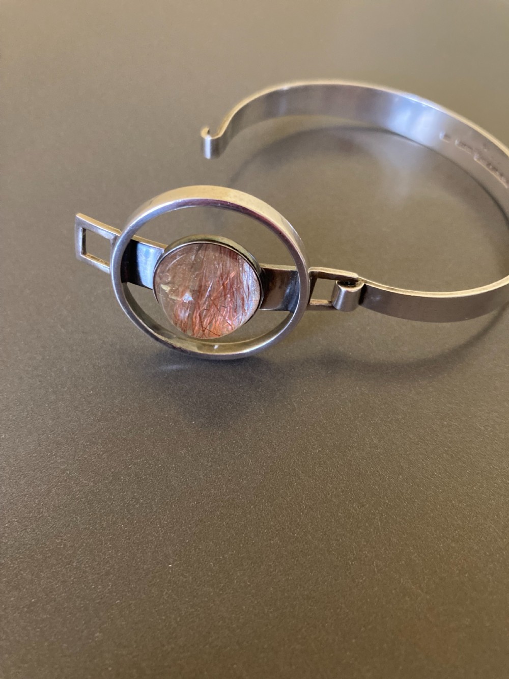 A hinged silver bangle features a central circular frame holding a cabochon rutilated quartz gemstone with needle-like inclusions. View IV