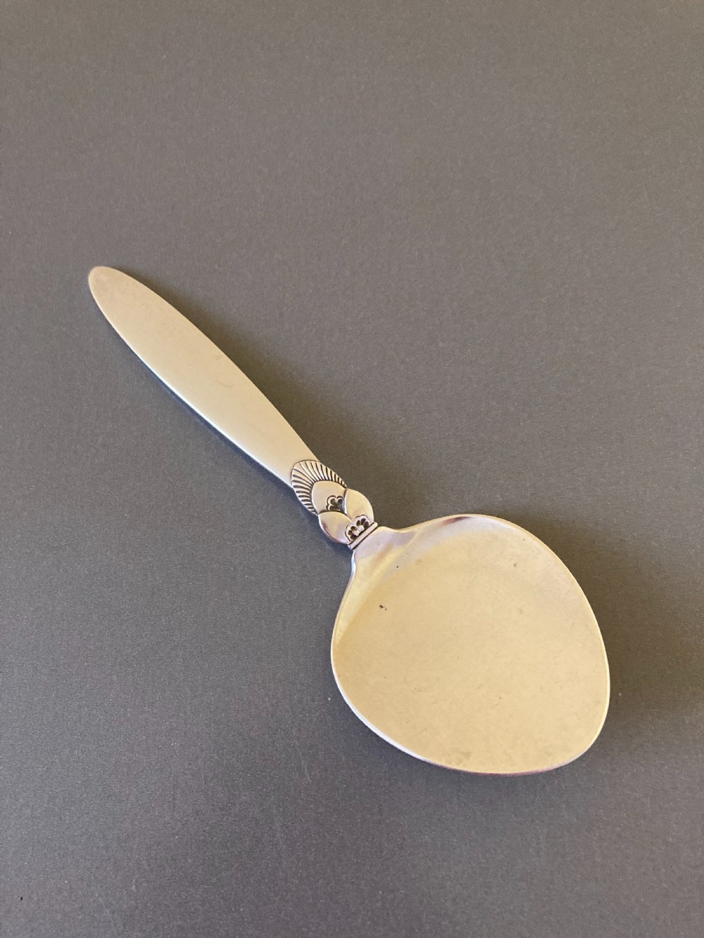 An overhead view of a vintage silver pastry spoon featuring an elongated handle and an embossed botanical motif near the wide bowl.