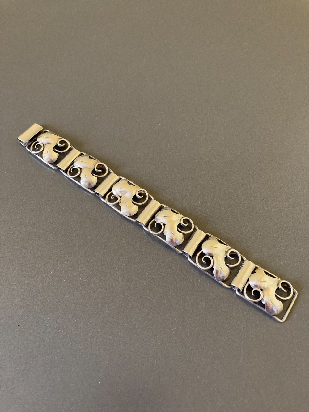 An overhead view of a linked silver bracelet featuring repeating organic scroll motifs against a dark grey background.