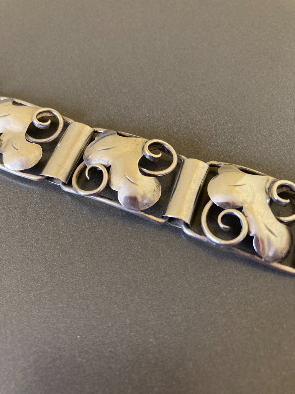 An overhead view of a linked silver bracelet featuring repeating organic scroll motifs against a dark grey background. View II