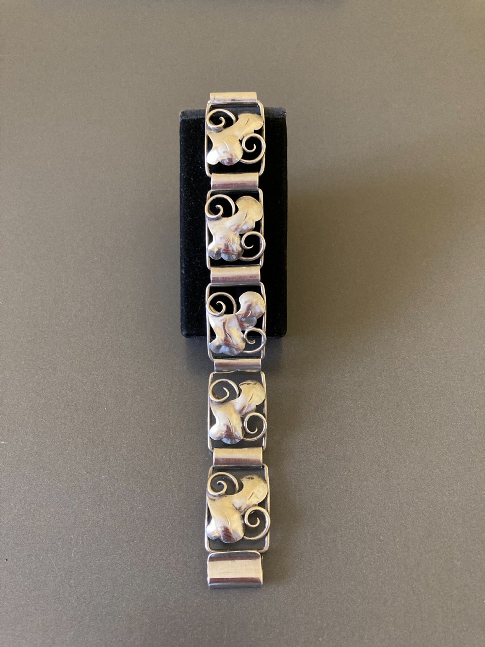 An overhead view of a linked silver bracelet featuring repeating organic scroll motifs against a dark grey background. View III