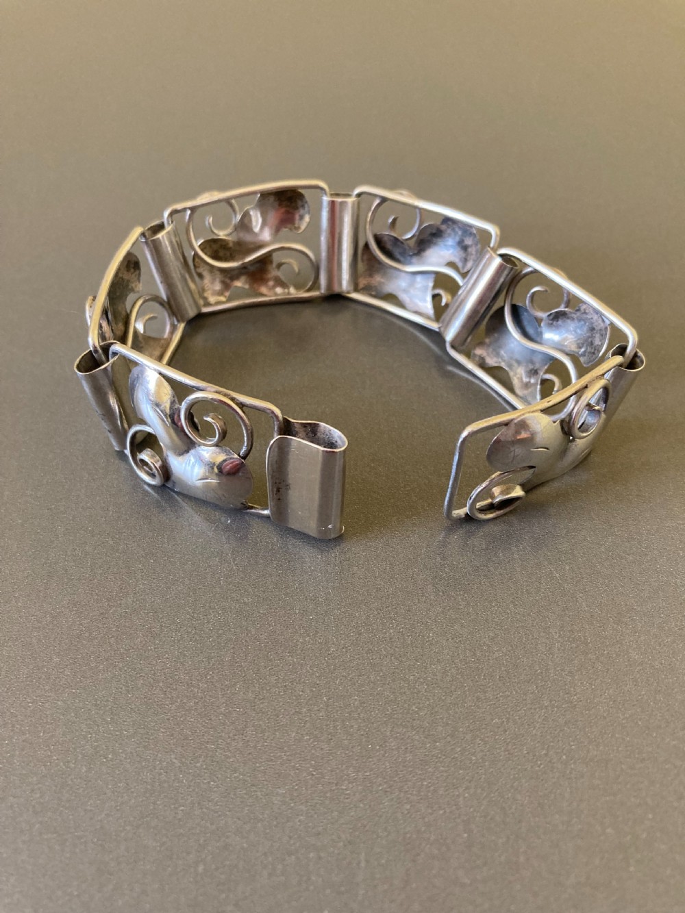 An overhead view of a linked silver bracelet featuring repeating organic scroll motifs against a dark grey background. View IV