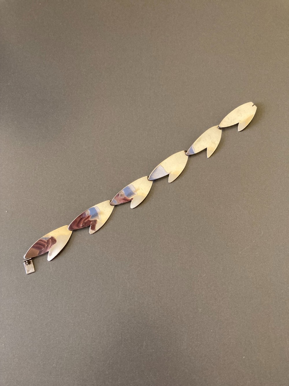 An overhead view of a linked silver bracelet featuring geometric petal-shaped components arranged in a linear sequence on a neutral grey background.