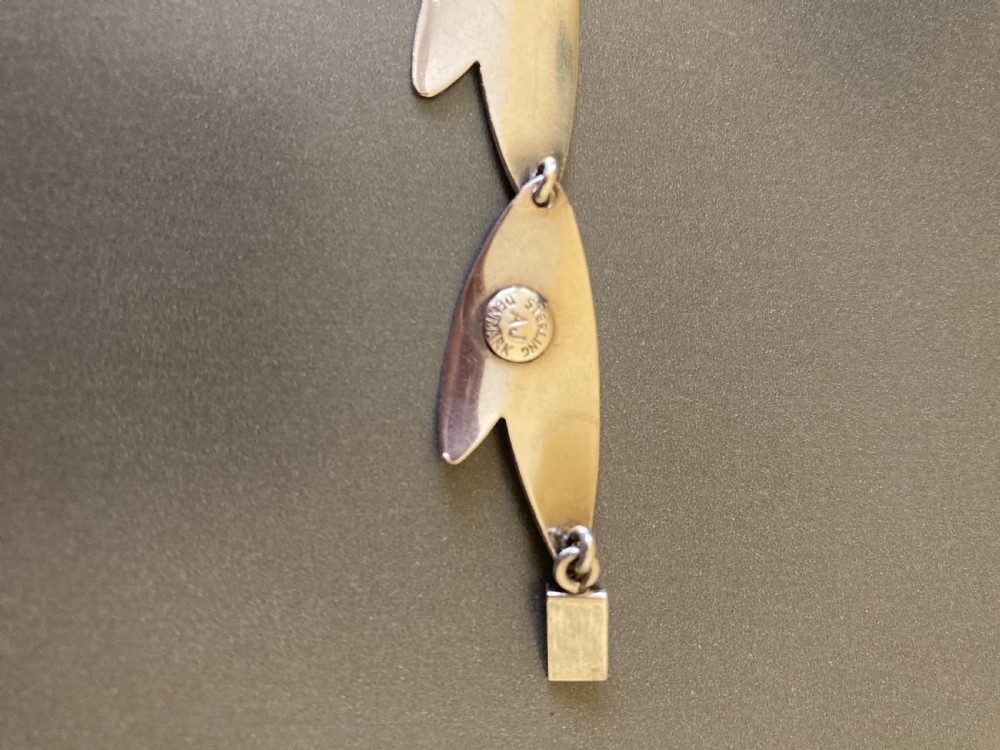 An overhead view of a linked silver bracelet featuring geometric petal-shaped components arranged in a linear sequence on a neutral grey background. View III