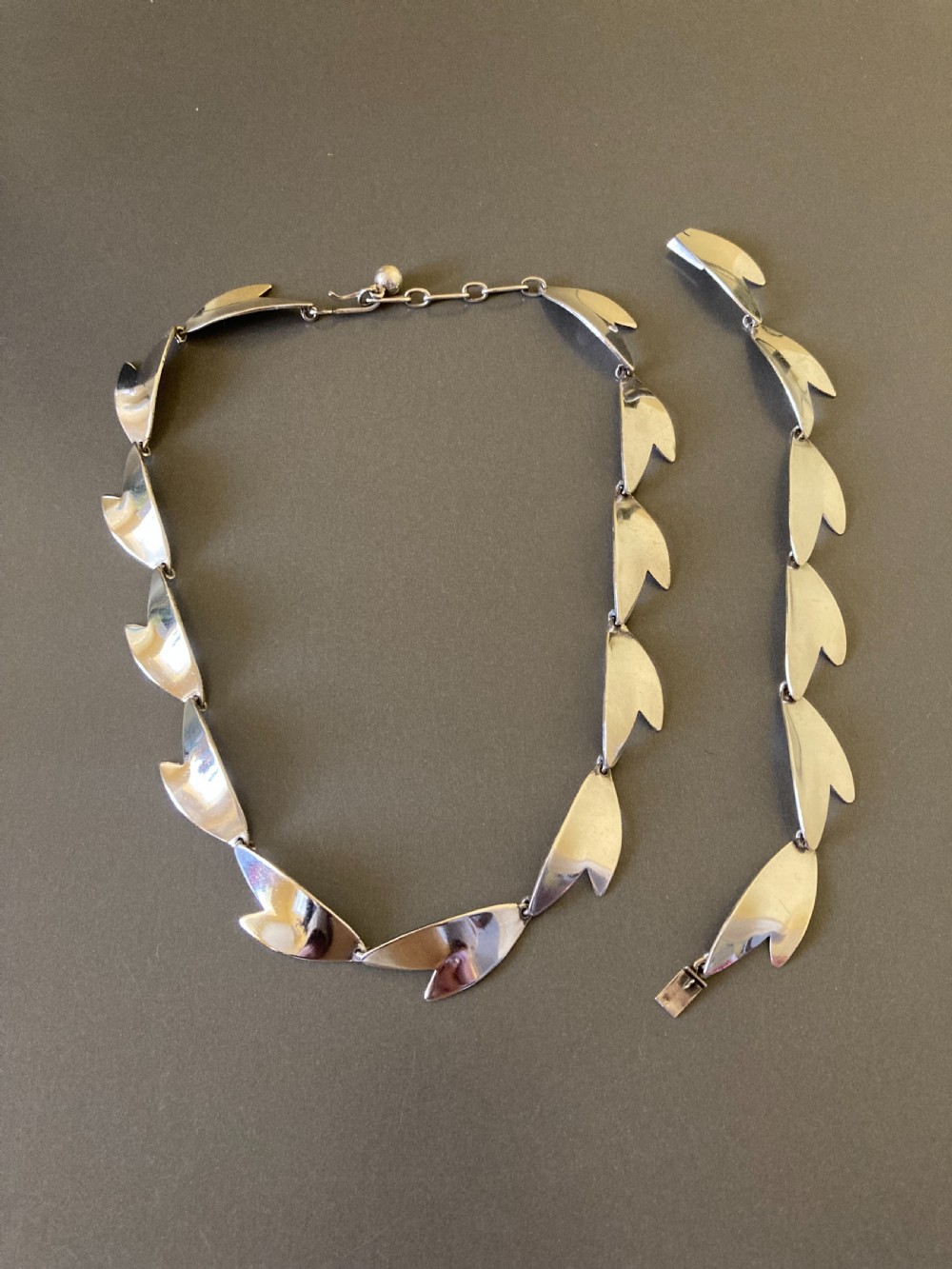 An overhead view of a linked silver bracelet featuring geometric petal-shaped components arranged in a linear sequence on a neutral grey background. View V