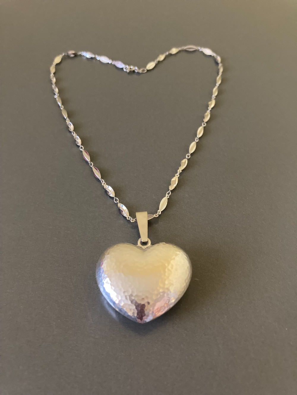 A hammered silver heart pendant suspended from a patterned sterling silver chain on a dark grey surface.