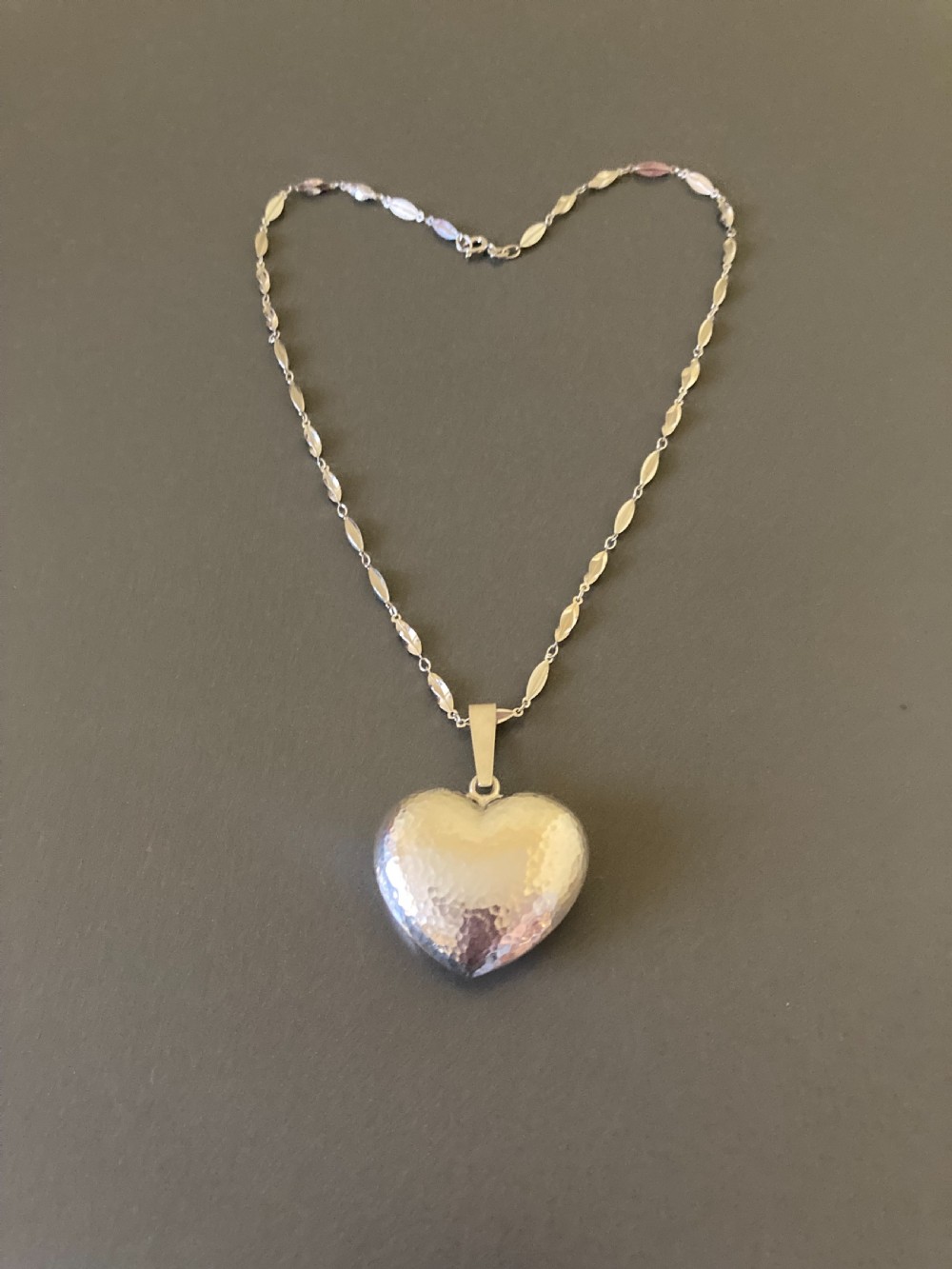 A hammered silver heart pendant suspended from a patterned sterling silver chain on a dark grey surface. View III