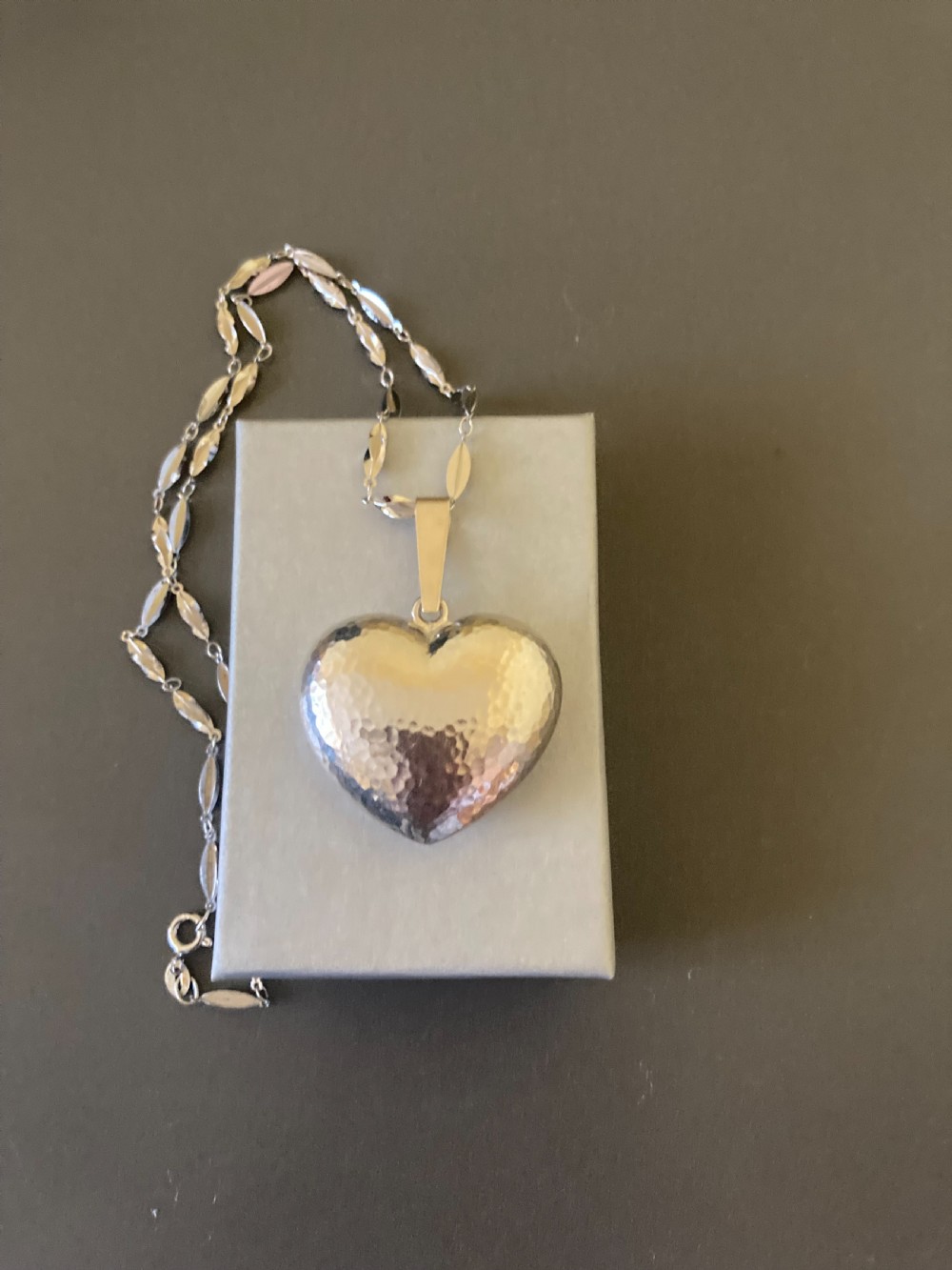 A hammered silver heart pendant suspended from a patterned sterling silver chain on a dark grey surface. View VI