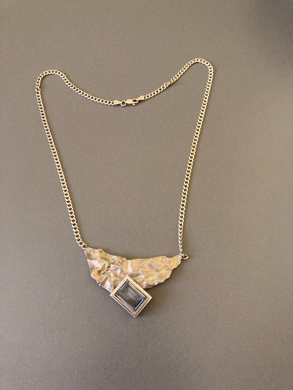 A 1960s sterling silver curb chain necklace featuring a textured asymmetrical pendant with an emerald-cut blue topaz stone.