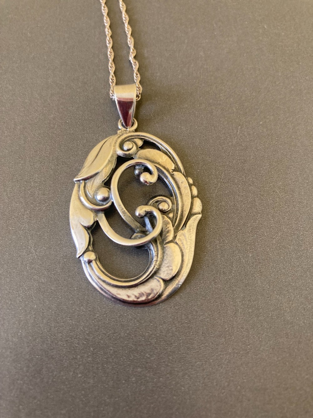 An oval silver pendant featuring openwork scroll details and leaf motifs suspended from a fine silver chain.