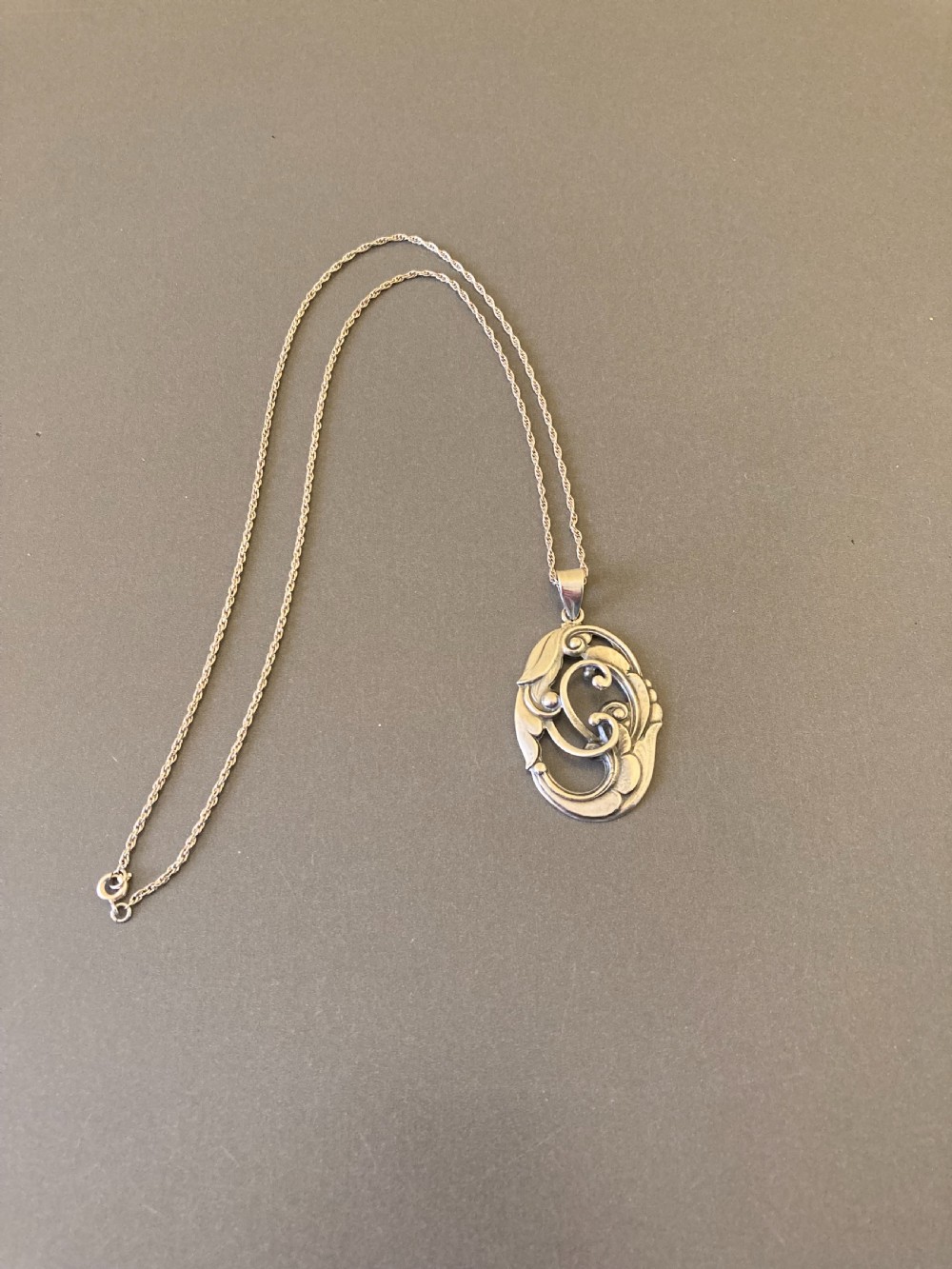An oval silver pendant featuring openwork scroll details and leaf motifs suspended from a fine silver chain. View II