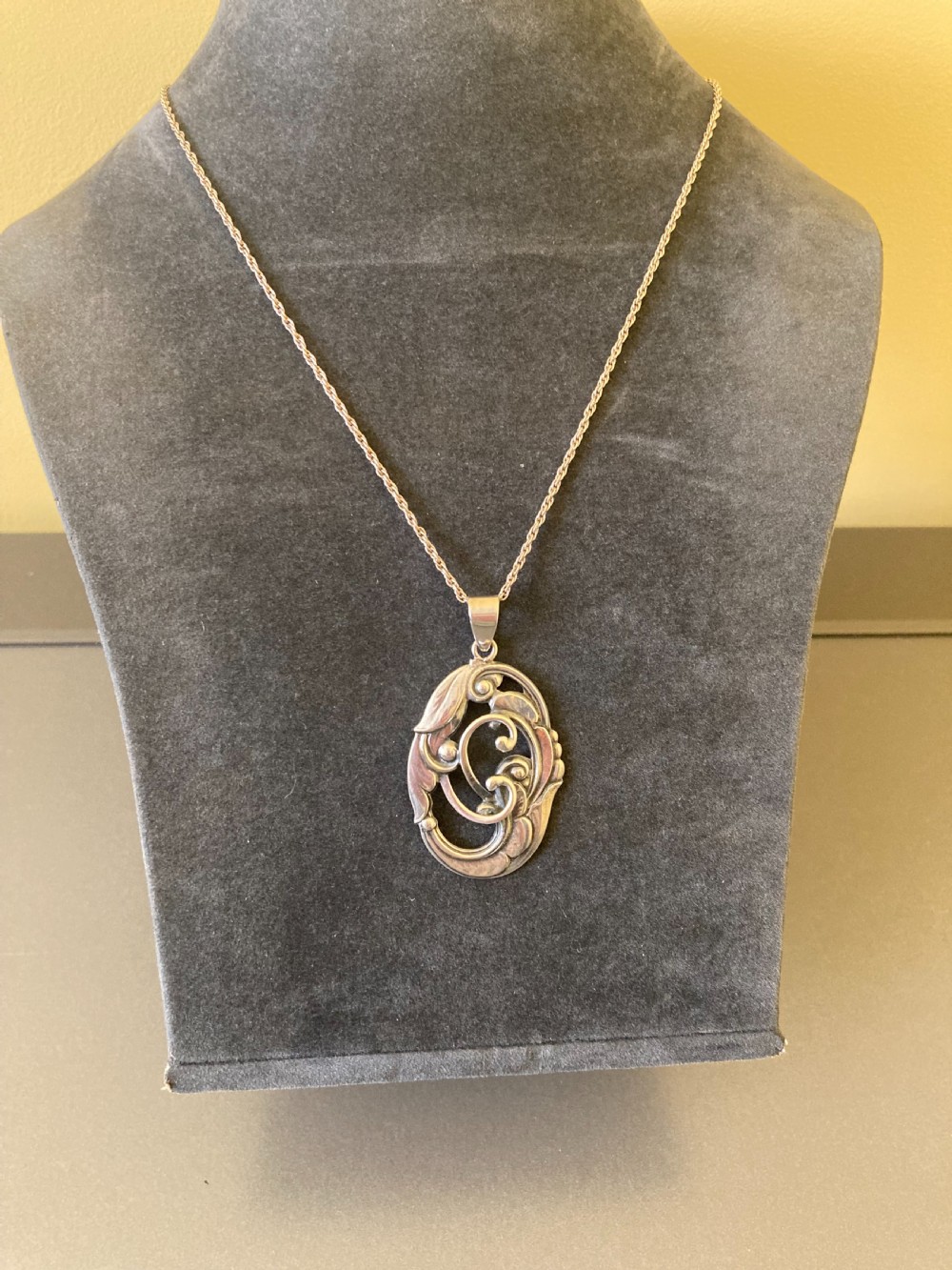 An oval silver pendant featuring openwork scroll details and leaf motifs suspended from a fine silver chain. View III