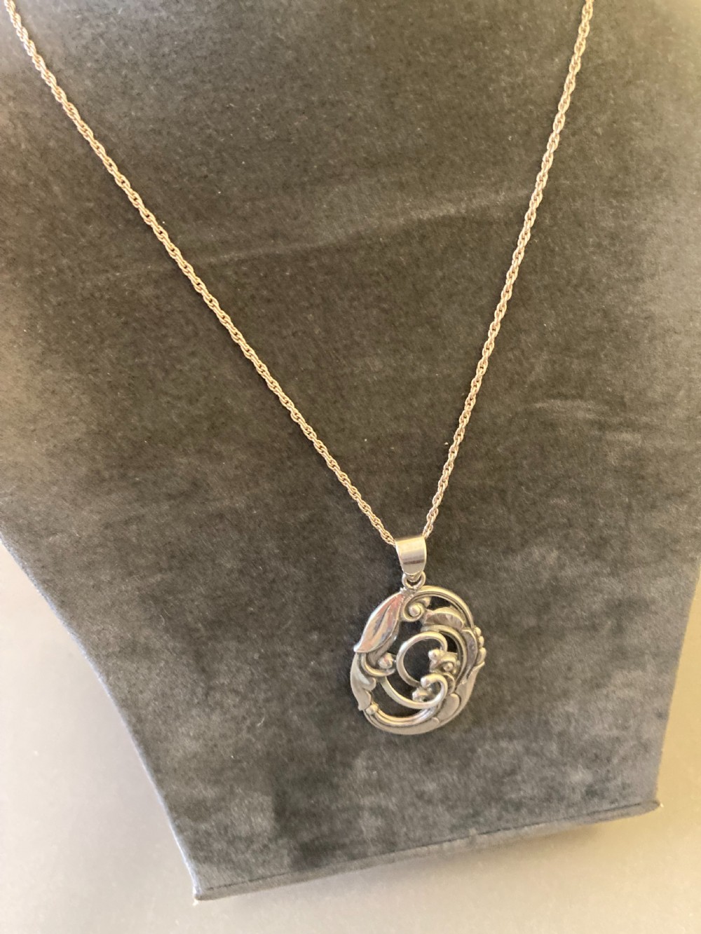 An oval silver pendant featuring openwork scroll details and leaf motifs suspended from a fine silver chain. View IV