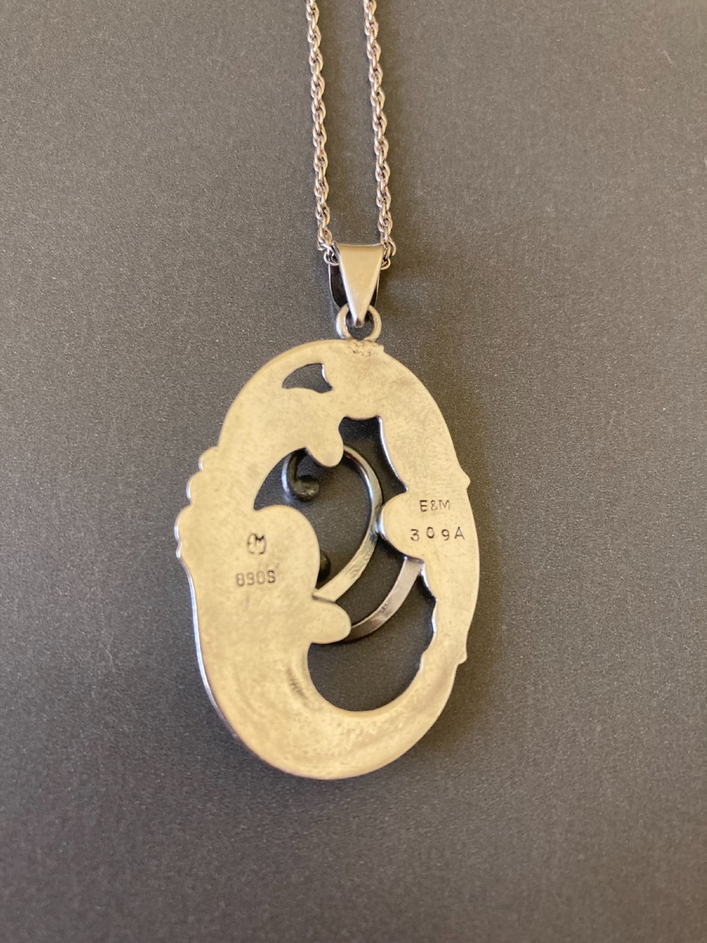 An oval silver pendant featuring openwork scroll details and leaf motifs suspended from a fine silver chain. View V