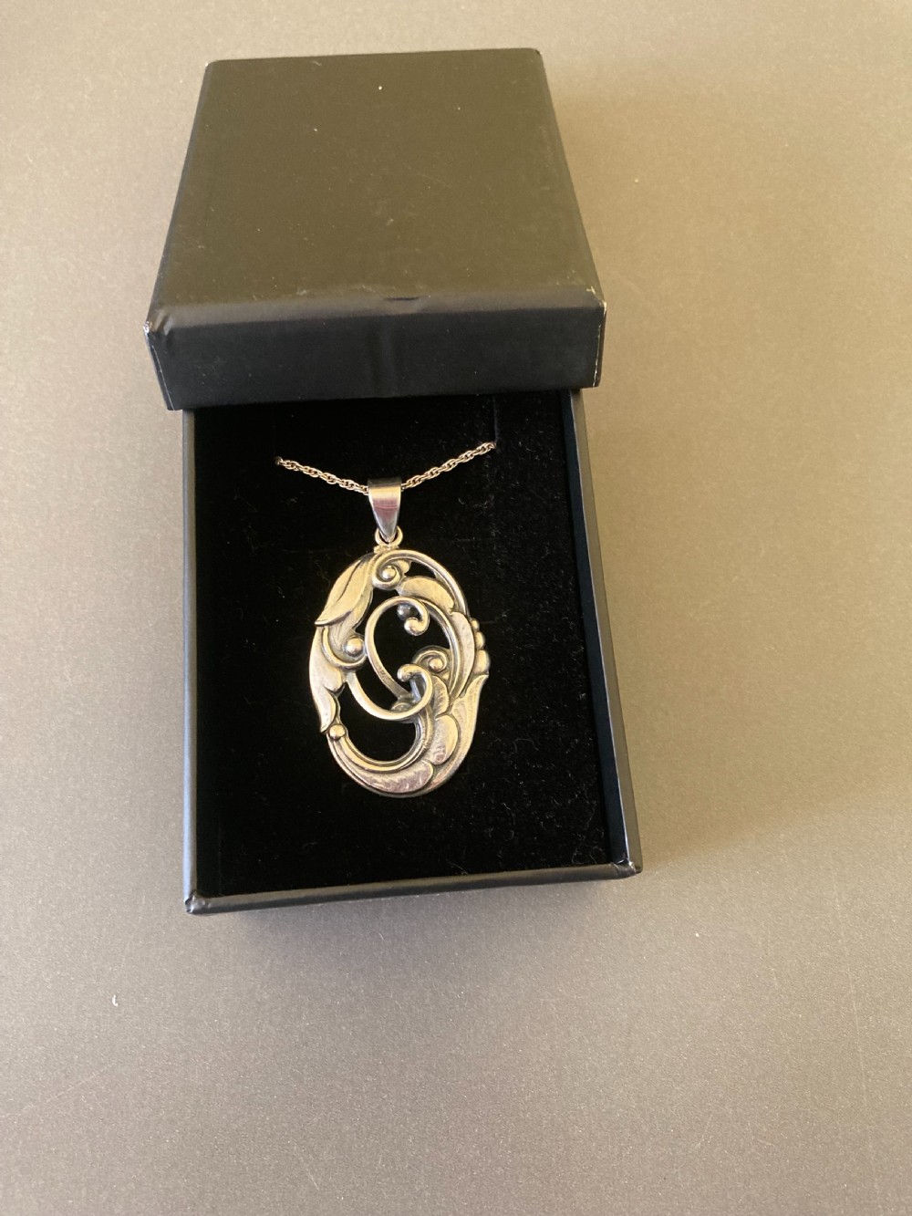 An oval silver pendant featuring openwork scroll details and leaf motifs suspended from a fine silver chain. View VI