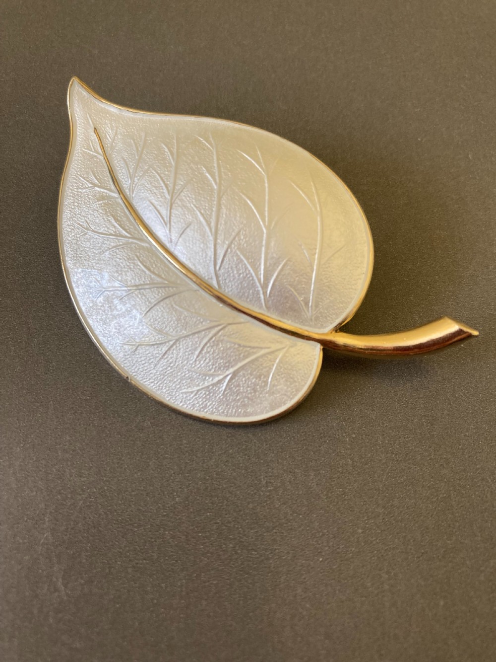 A leaf-shaped white guilloche enamel brooch with silver gilt edges and a curved stem.