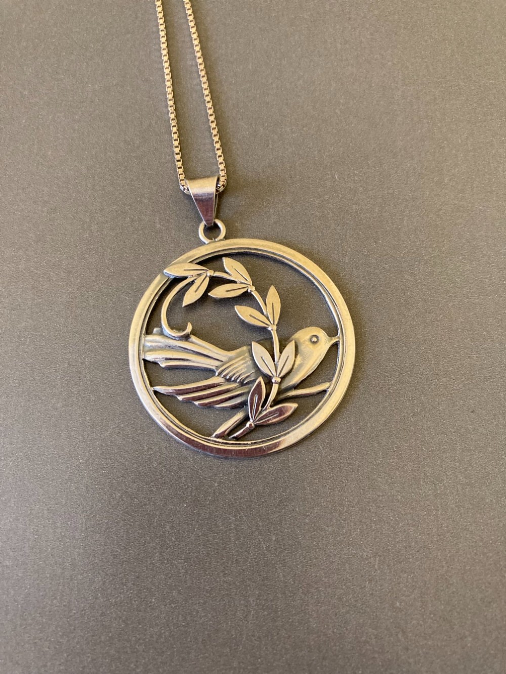 A circular silver pendant depicting a bird perched among leaf motifs within a round frame, suspended from a silver box chain.