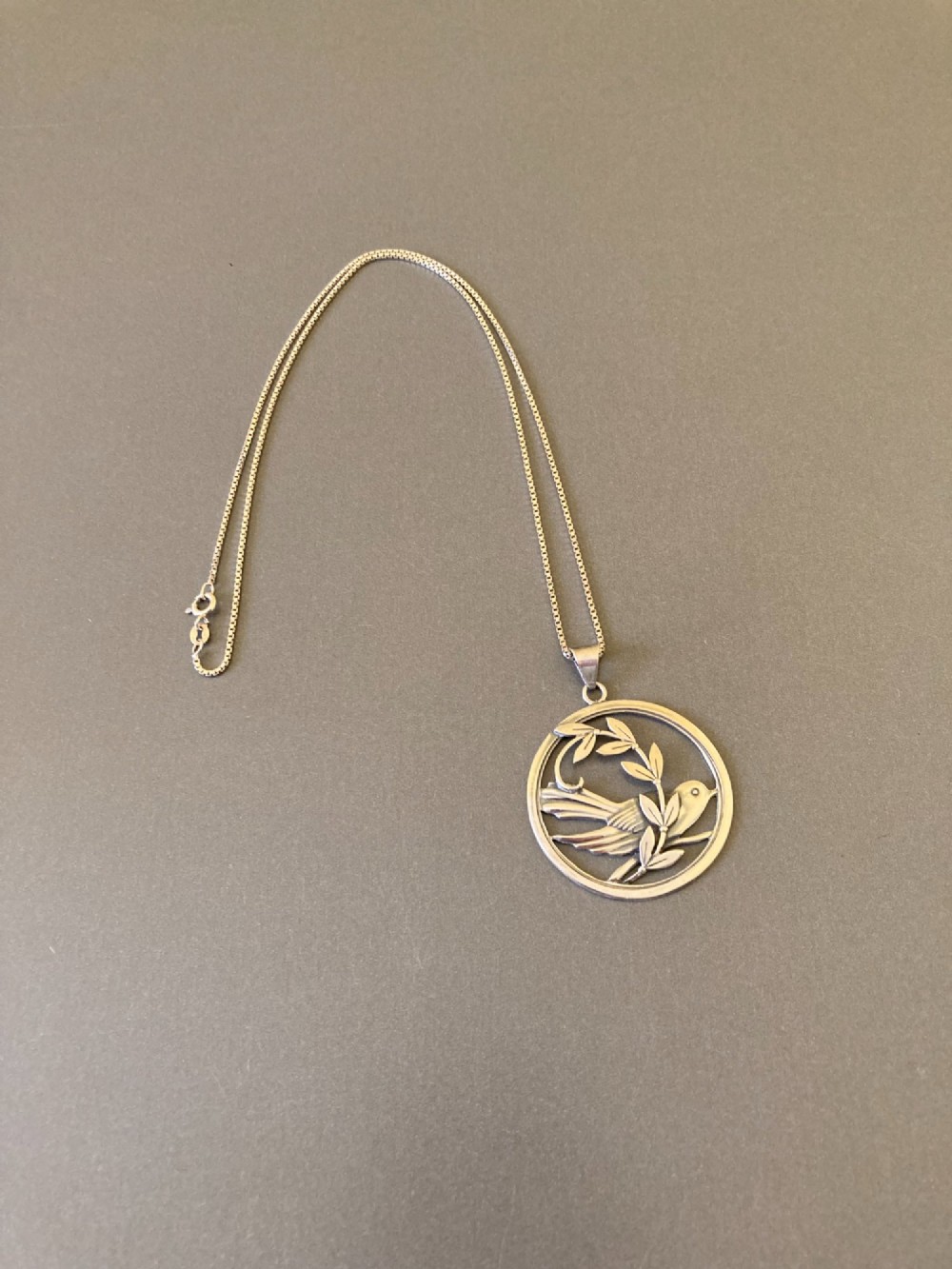 A circular silver pendant depicting a bird perched among leaf motifs within a round frame, suspended from a silver box chain. View II