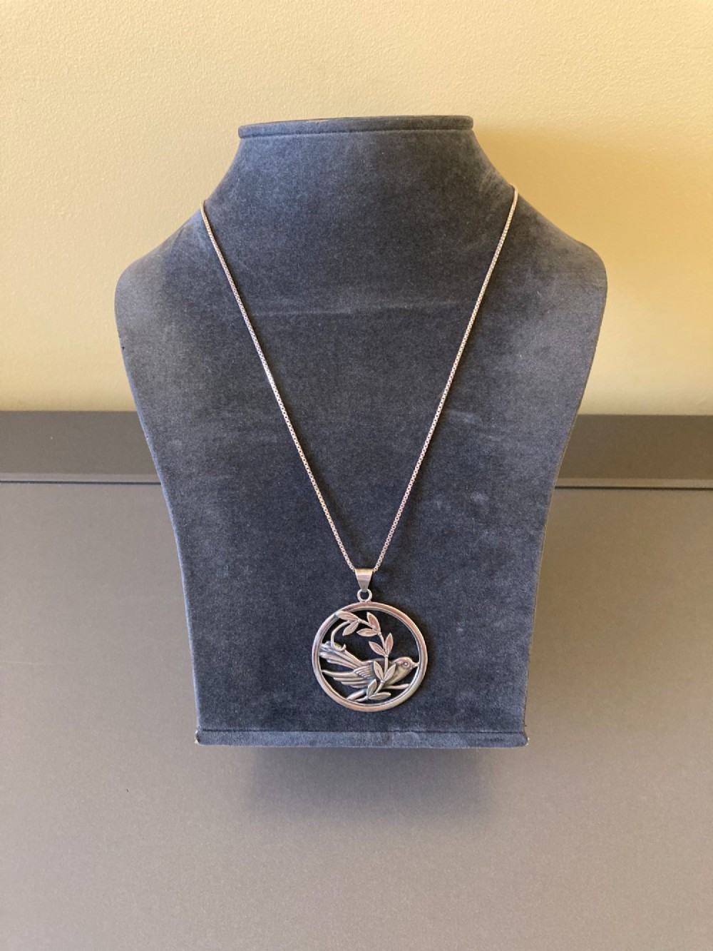 A circular silver pendant depicting a bird perched among leaf motifs within a round frame, suspended from a silver box chain. View III