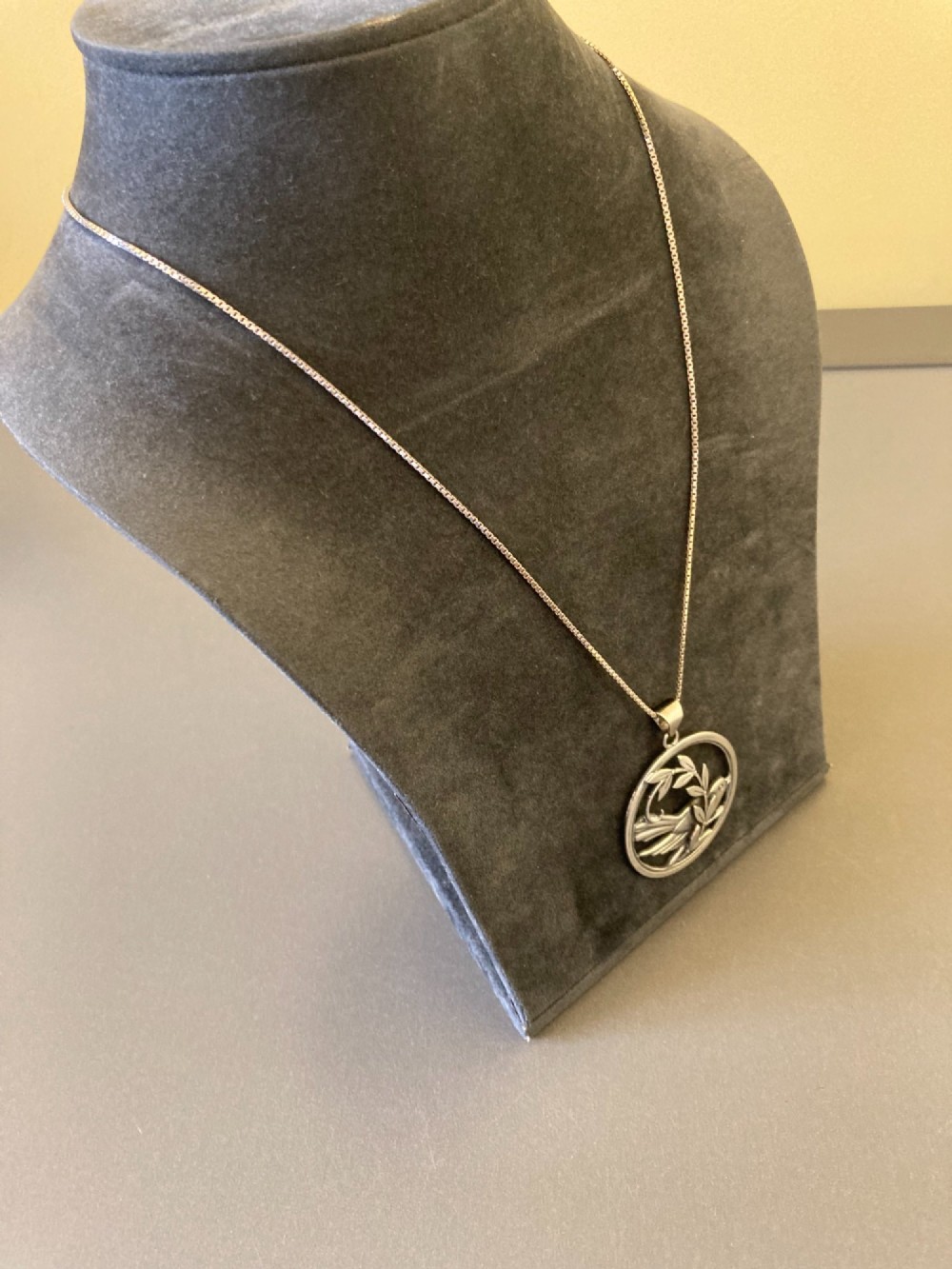 A circular silver pendant depicting a bird perched among leaf motifs within a round frame, suspended from a silver box chain. View IV