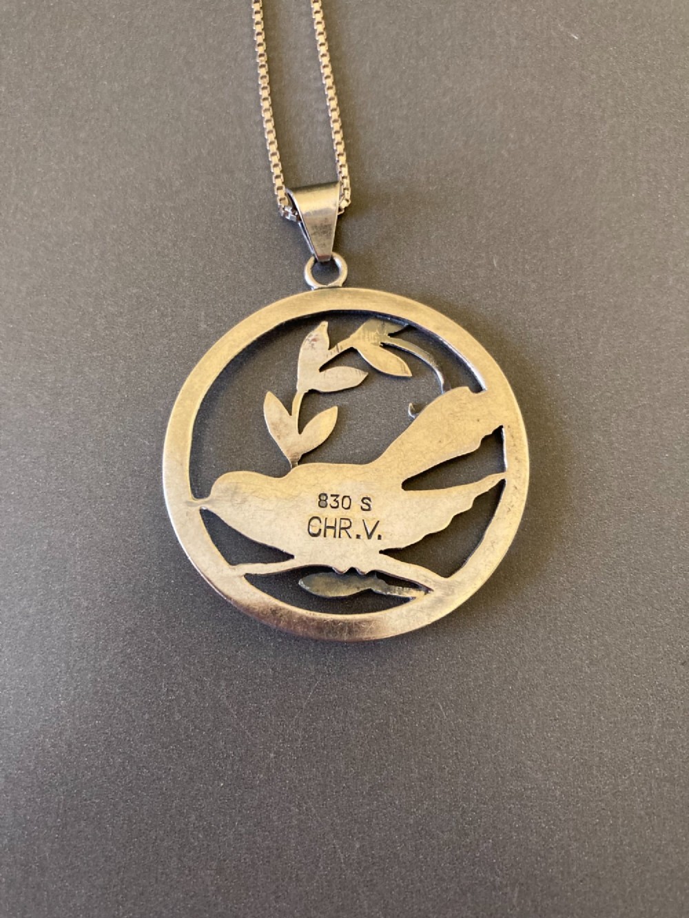 A circular silver pendant depicting a bird perched among leaf motifs within a round frame, suspended from a silver box chain. View V
