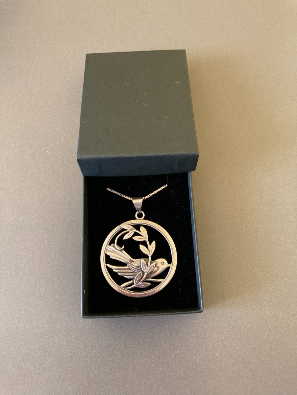 A circular silver pendant depicting a bird perched among leaf motifs within a round frame, suspended from a silver box chain. View VI