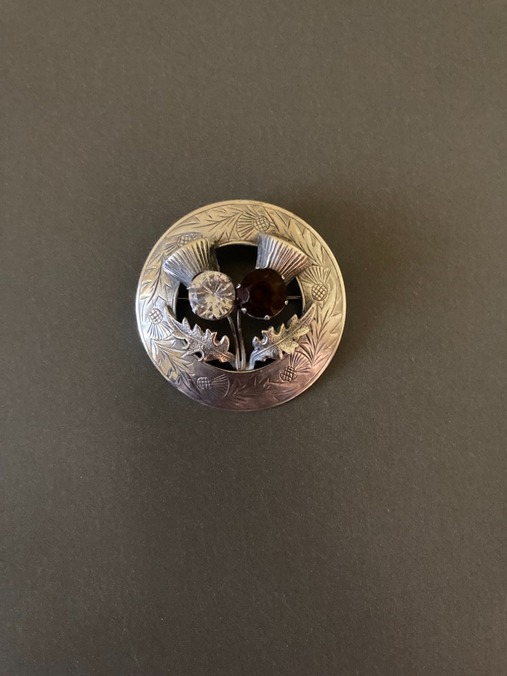 A circular silver brooch featuring hand-engraved leaf patterns, two detailed thistle heads, and two round faceted stones in clear and brown shades. View II