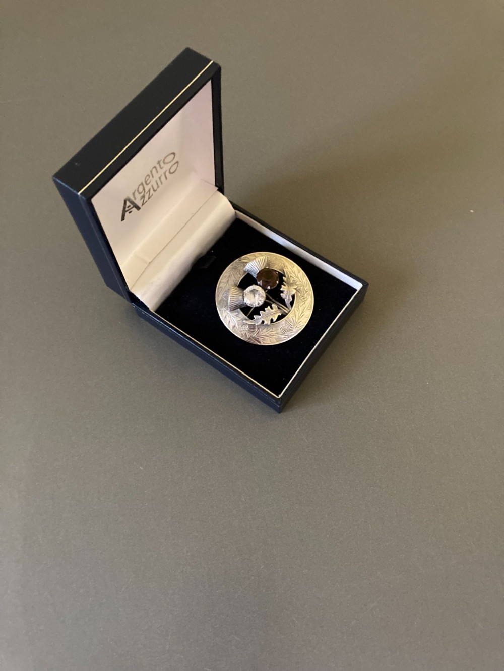 A circular silver brooch featuring hand-engraved leaf patterns, two detailed thistle heads, and two round faceted stones in clear and brown shades. View III