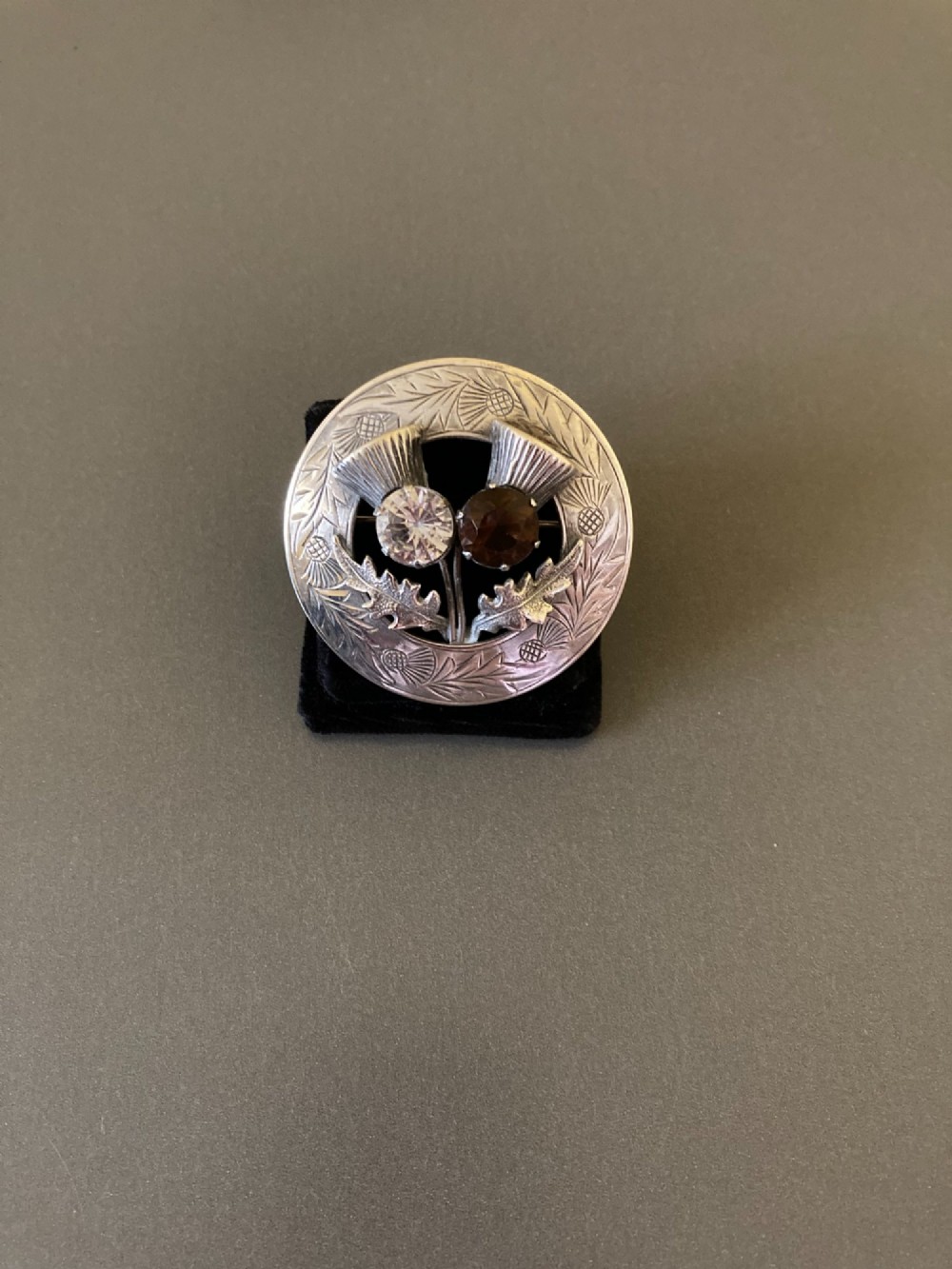 A circular silver brooch featuring hand-engraved leaf patterns, two detailed thistle heads, and two round faceted stones in clear and brown shades. View IV