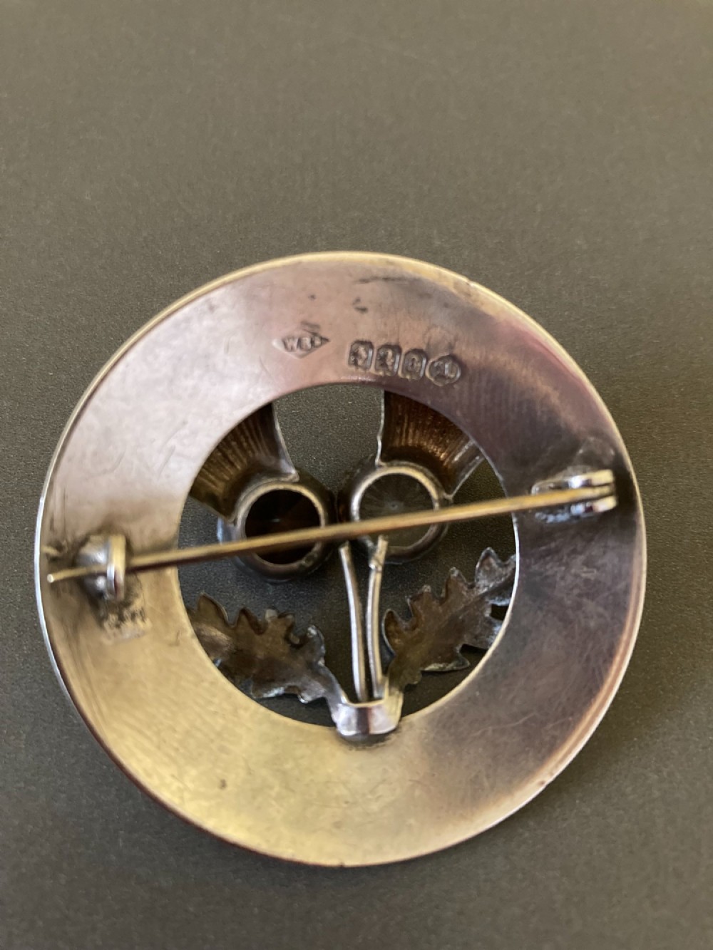 A circular silver brooch featuring hand-engraved leaf patterns, two detailed thistle heads, and two round faceted stones in clear and brown shades. View V