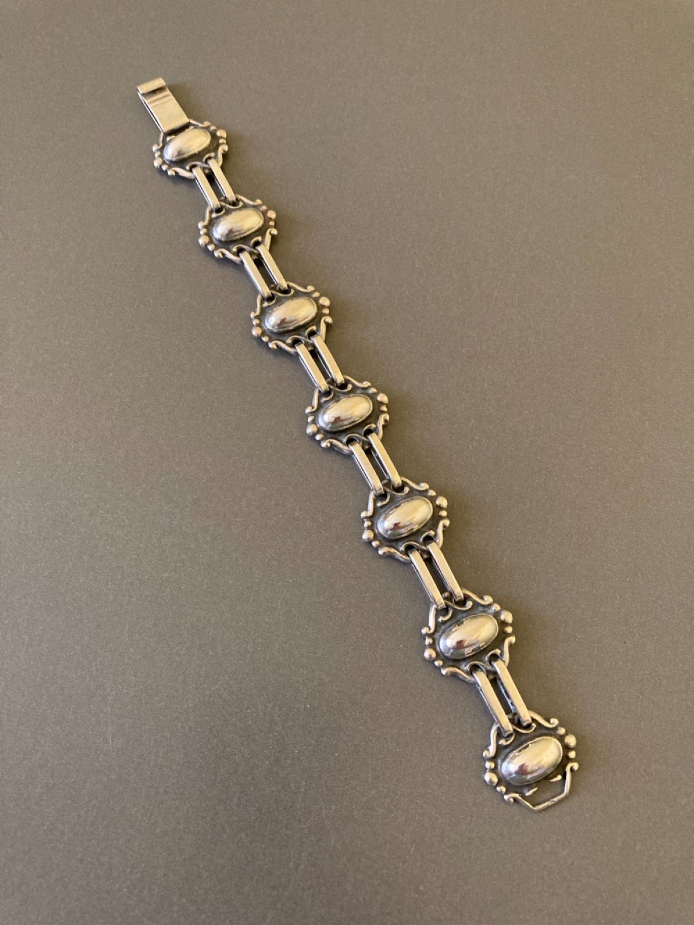 A 1930s Danish silver link bracelet featuring seven raised oval cabochon motifs surrounded by scrollwork borders and joined by parallel bar links.