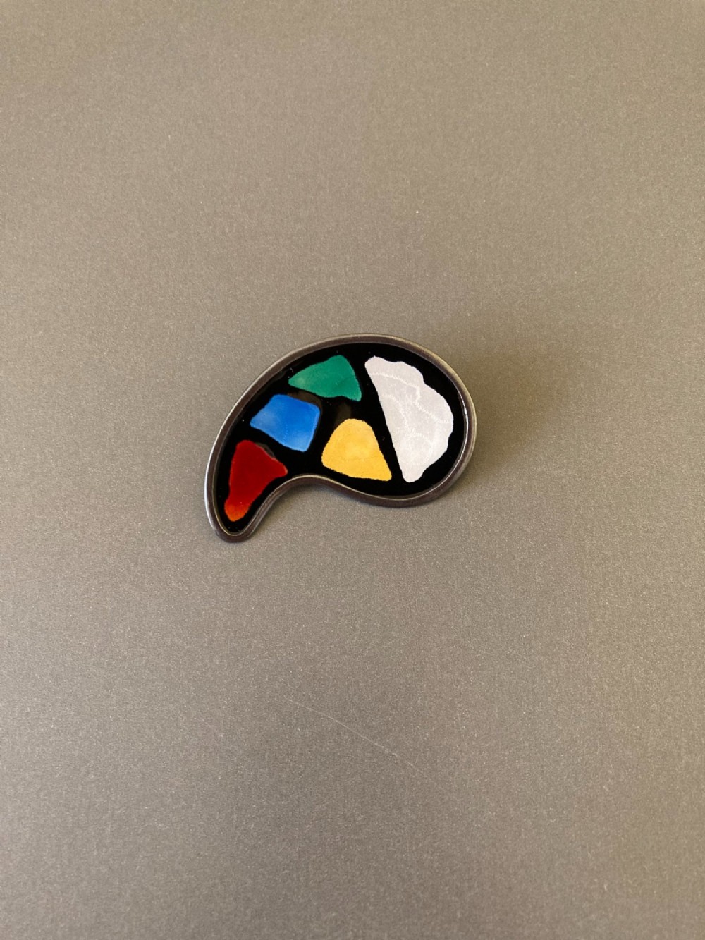 A silver brooch shaped like an artists palette with a black enamel background and five colourful enamel paint swatches.