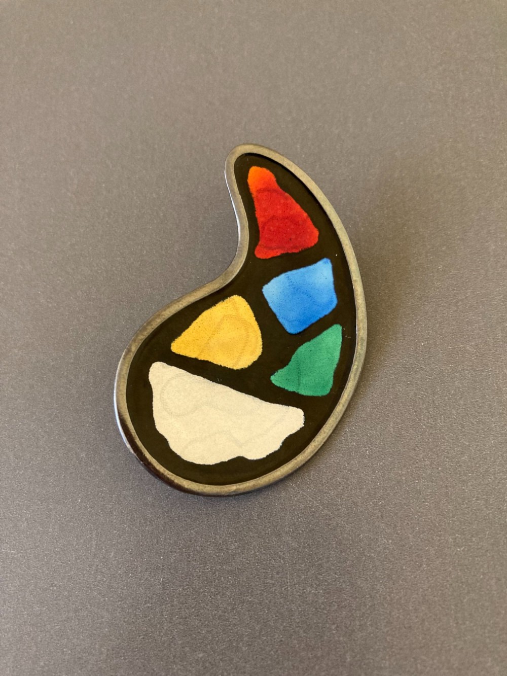 A silver brooch shaped like an artists palette with a black enamel background and five colourful enamel paint swatches. View II