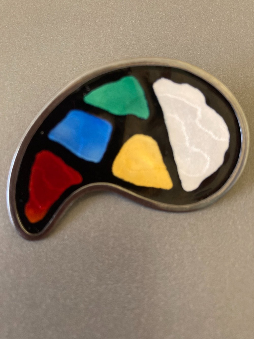 A silver brooch shaped like an artists palette with a black enamel background and five colourful enamel paint swatches. View V