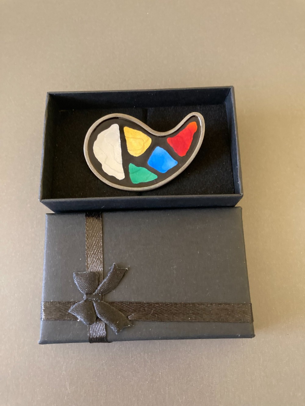 A silver brooch shaped like an artists palette with a black enamel background and five colourful enamel paint swatches. View VI
