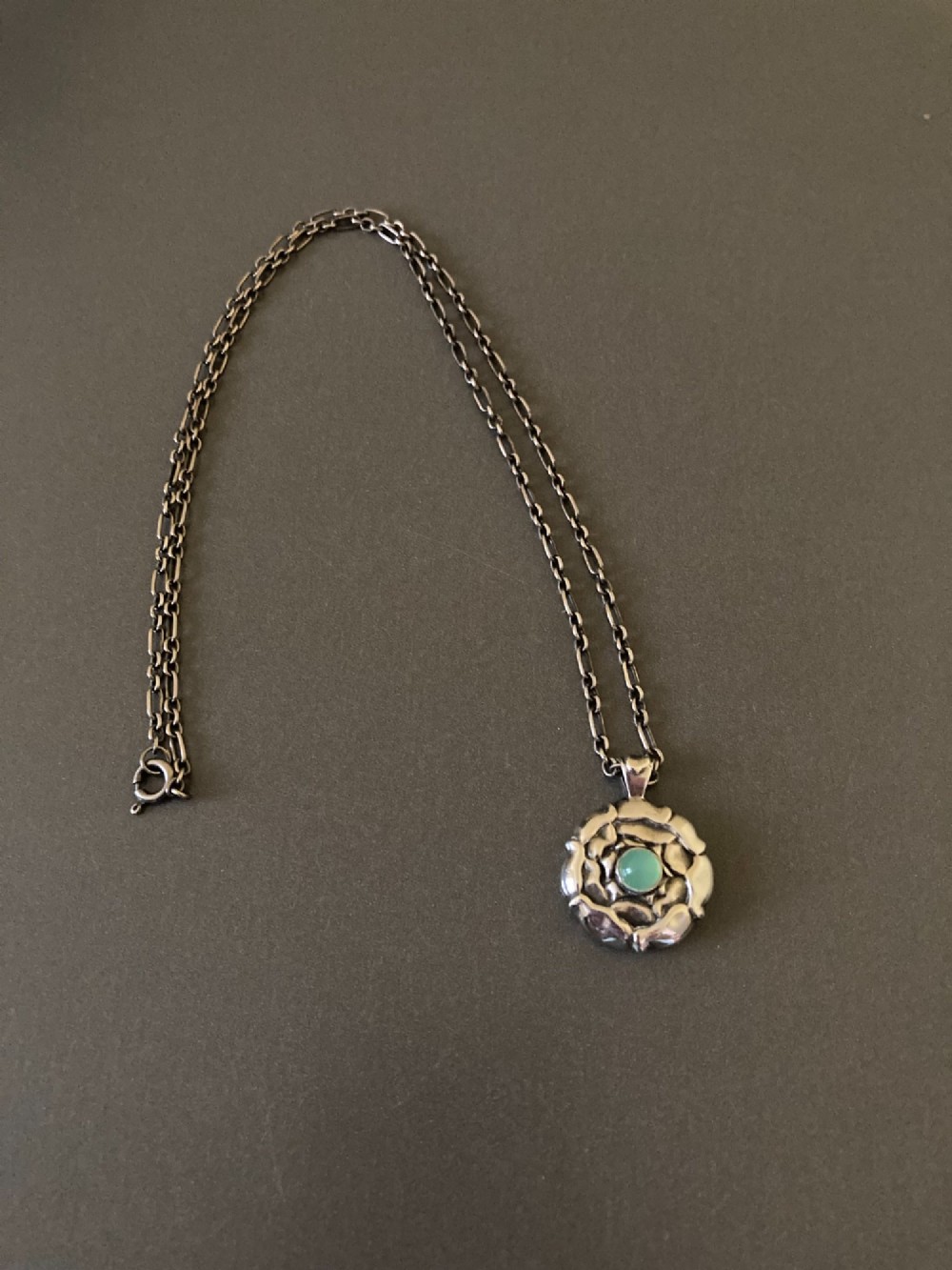 A circular sterling silver pendant with relief floral petal detailing and a central green cabochon chrysoprase, hanging from a silver link chain.