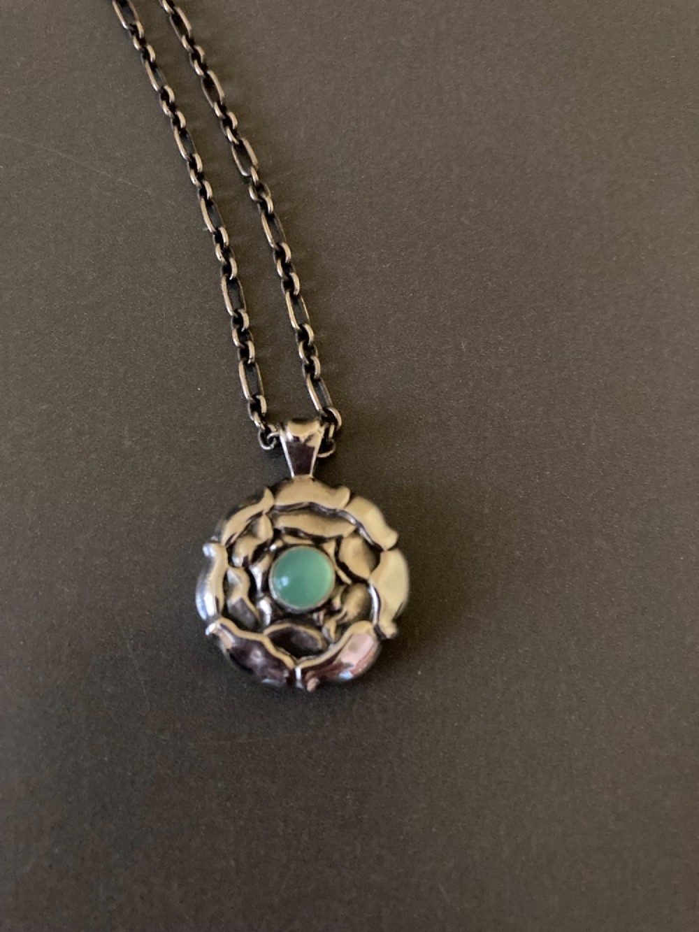 A circular sterling silver pendant with relief floral petal detailing and a central green cabochon chrysoprase, hanging from a silver link chain. View IV