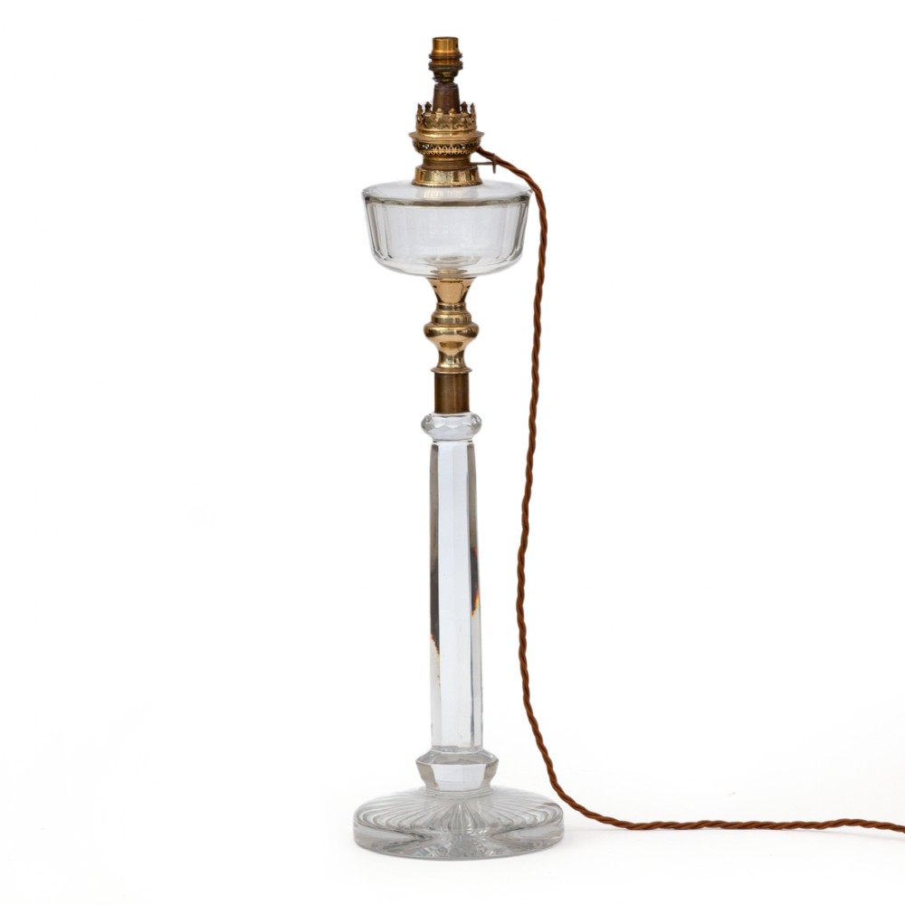 An antique lead crystal table lamp featuring a faceted glass column stem, cut glass reservoir, and brass fittings on a circular base.