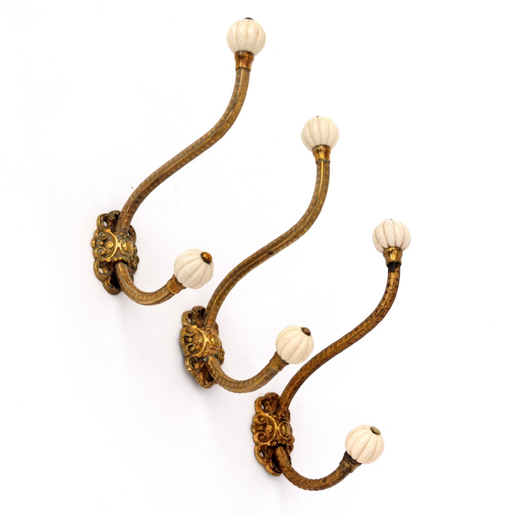 A set of three chased brass wall hooks with ornate scrollwork bases, S-curved stems, and fluted cream porcelain knobs.