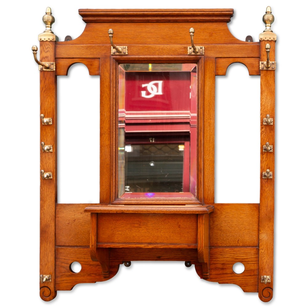 A wall-mounted golden oak coat rack featuring brass hooks, brass finials, a central bevelled mirror, and a small central storage box.