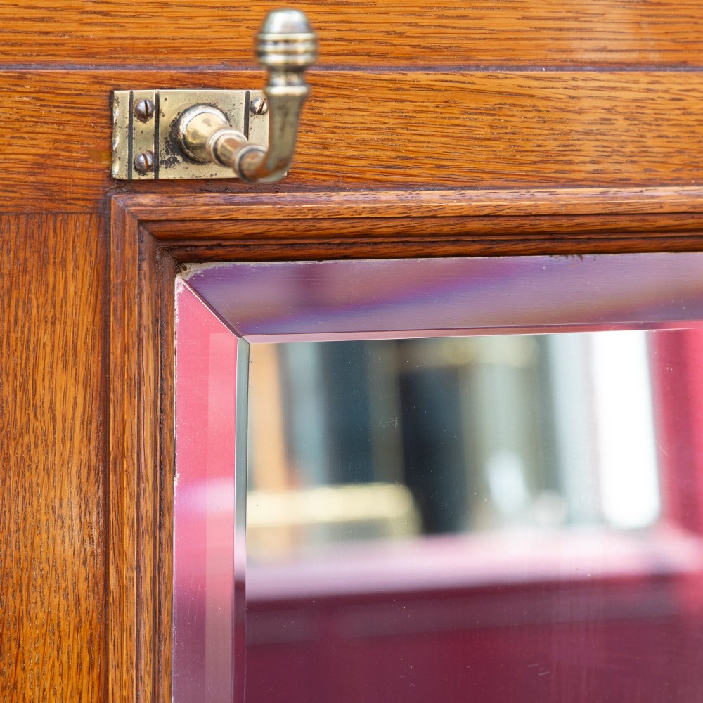 A wall-mounted golden oak coat rack featuring brass hooks, brass finials, a central bevelled mirror, and a small central storage box. View III