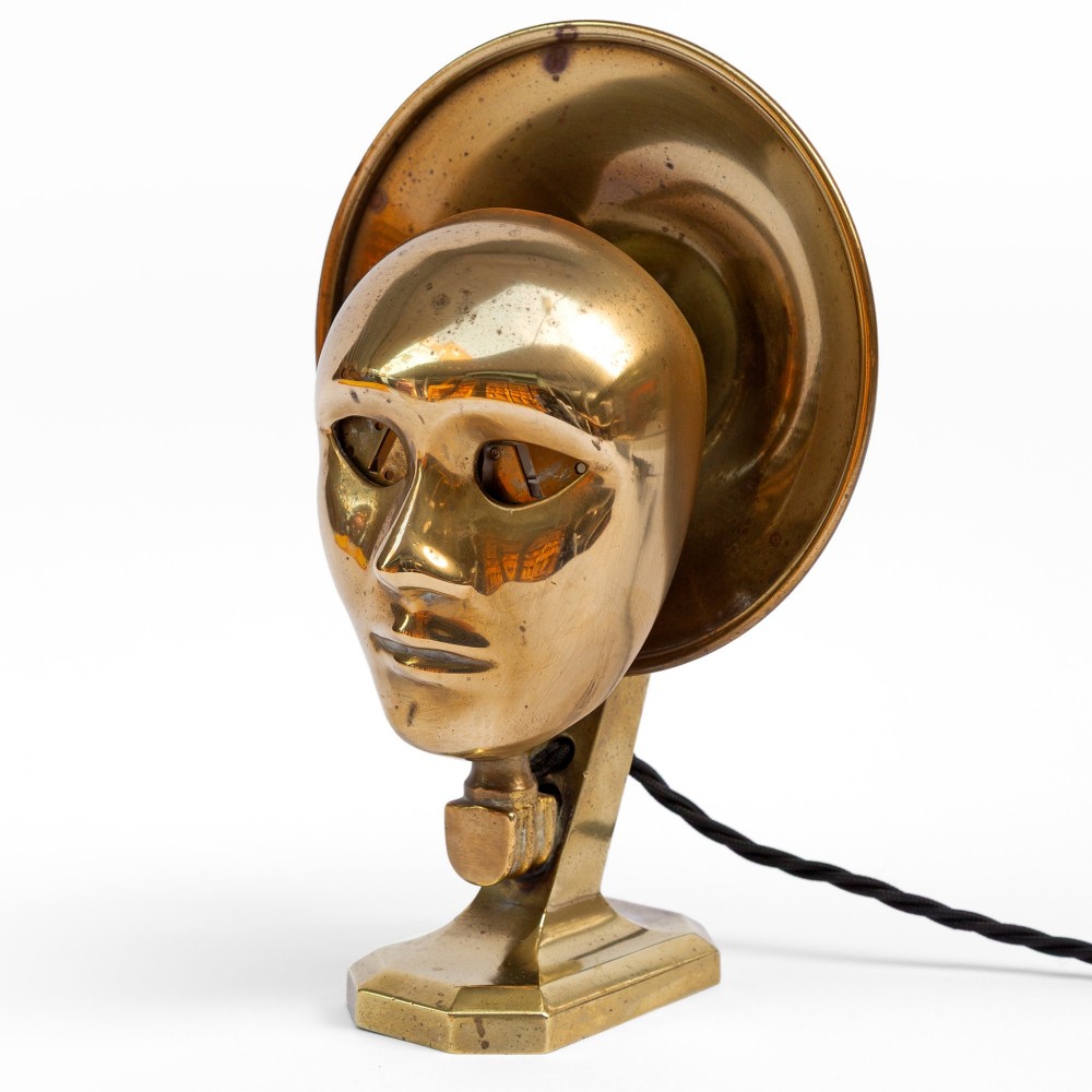 A polished brass table lamp featuring a stylised human-machine mask mounted in front of a circular dish reflector base.