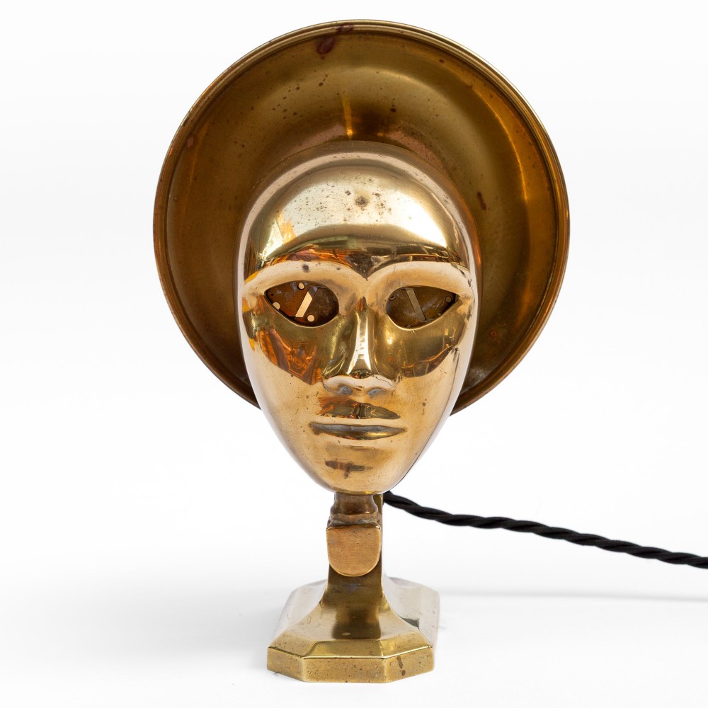 A polished brass table lamp featuring a stylised human-machine mask mounted in front of a circular dish reflector base. View II
