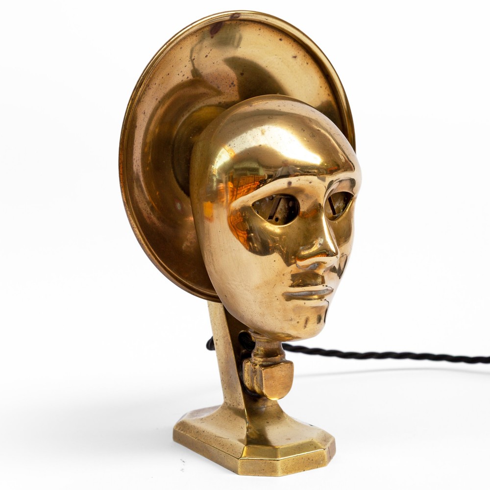 A polished brass table lamp featuring a stylised human-machine mask mounted in front of a circular dish reflector base. View III