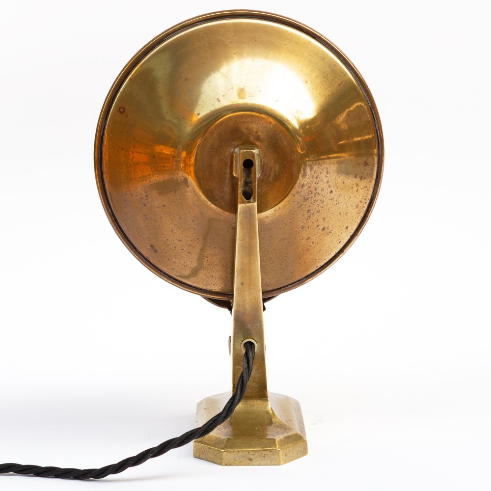 A polished brass table lamp featuring a stylised human-machine mask mounted in front of a circular dish reflector base. View IV