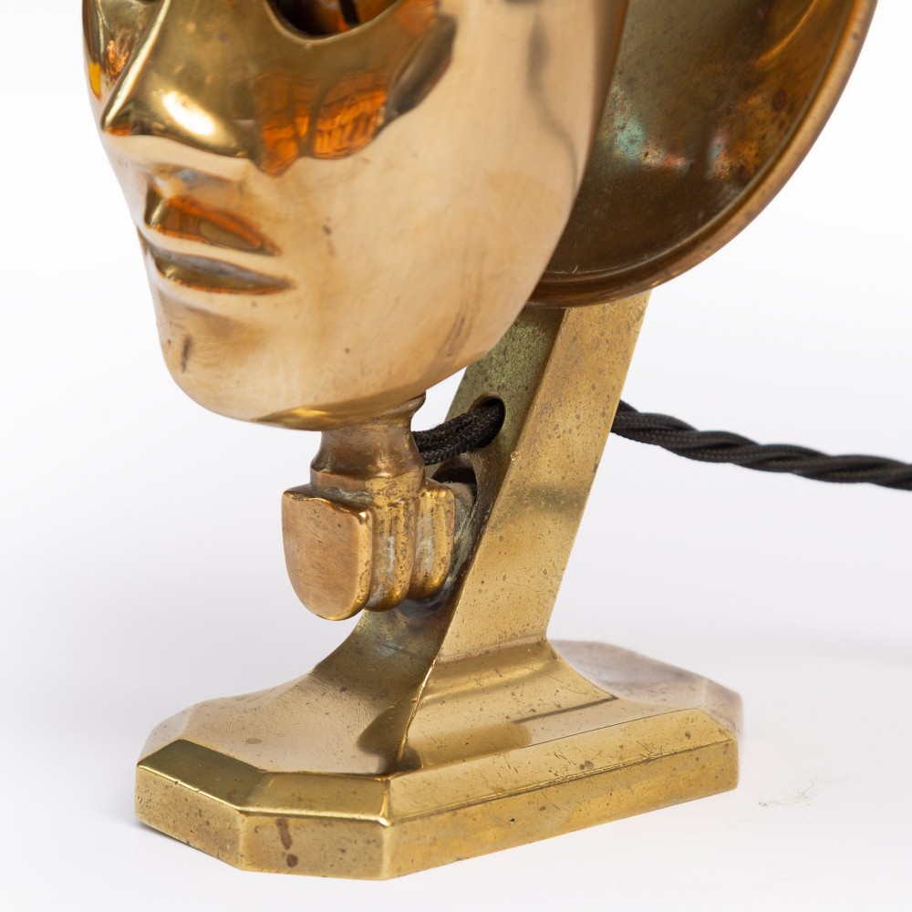 A polished brass table lamp featuring a stylised human-machine mask mounted in front of a circular dish reflector base. View V