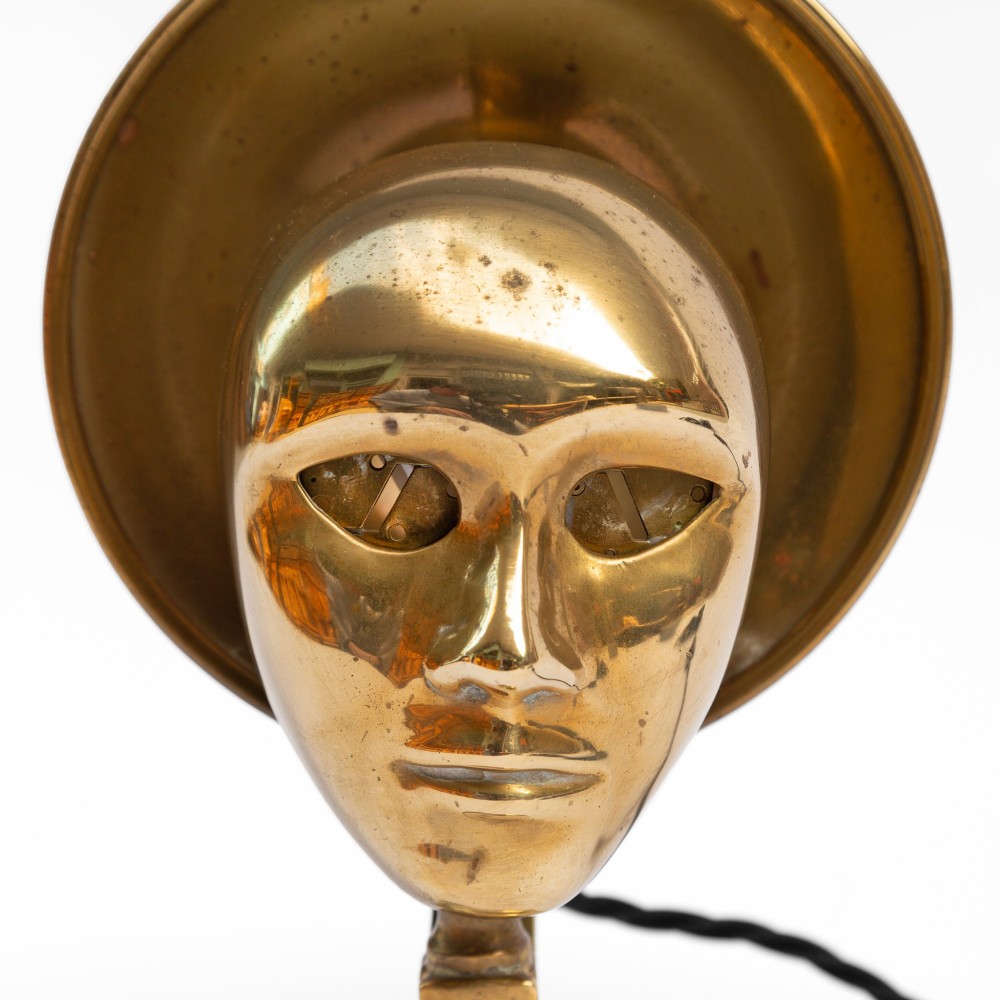 A polished brass table lamp featuring a stylised human-machine mask mounted in front of a circular dish reflector base. View VI