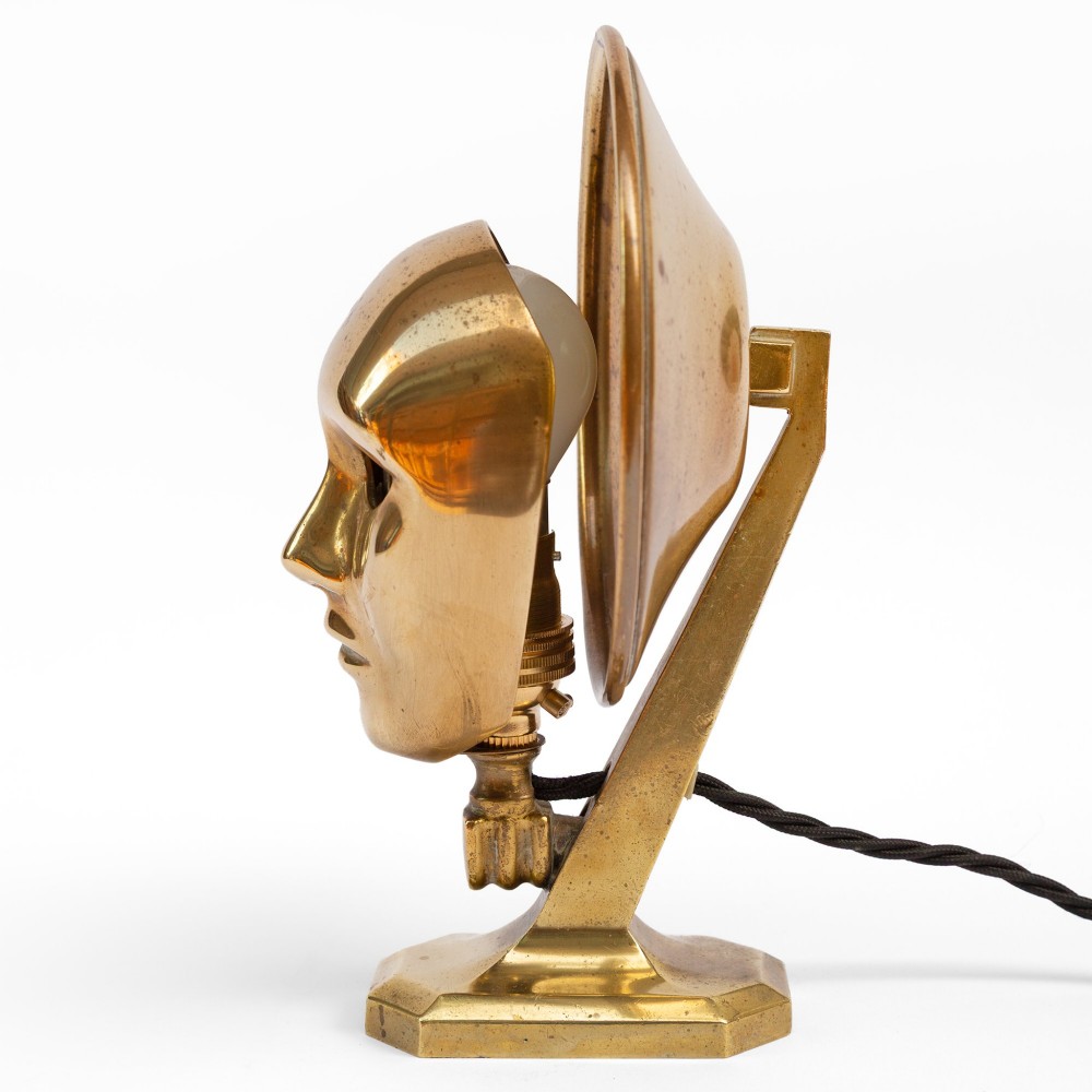 A polished brass table lamp featuring a stylised human-machine mask mounted in front of a circular dish reflector base. View VII