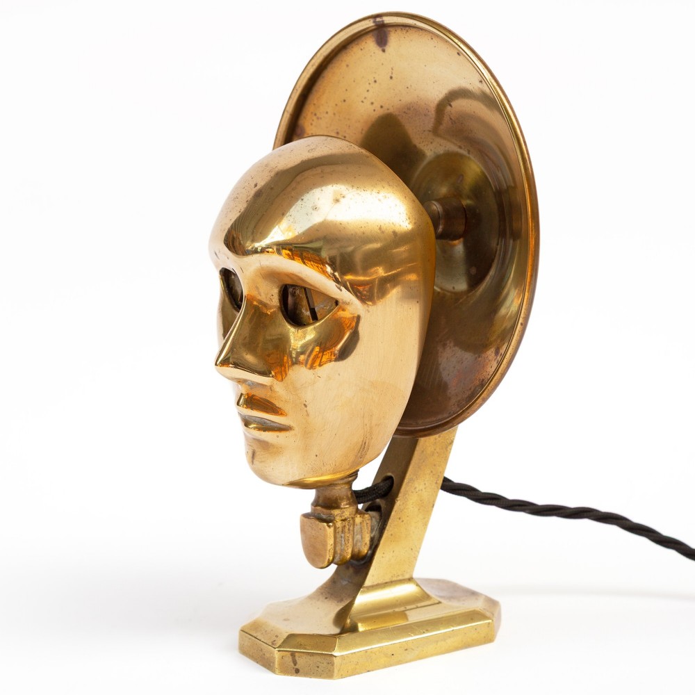 A polished brass table lamp featuring a stylised human-machine mask mounted in front of a circular dish reflector base. View VIII